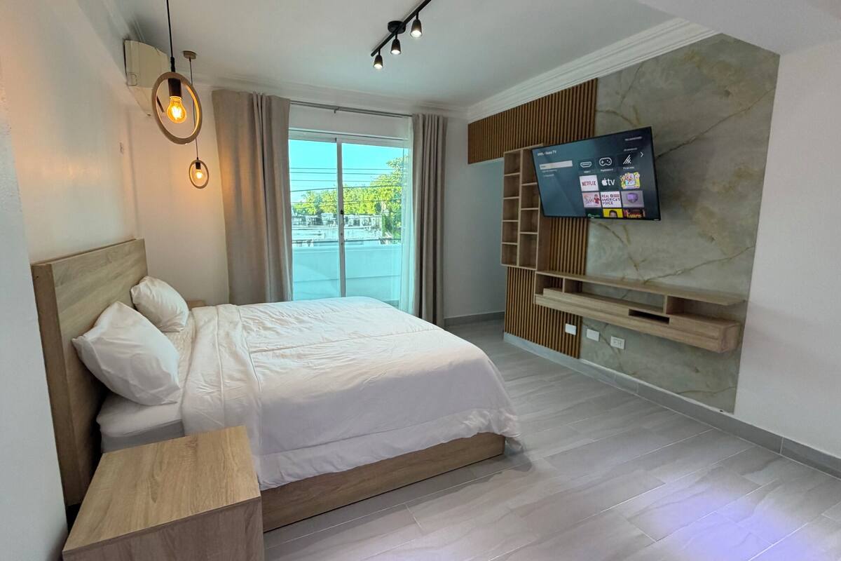 A modern hotel room with a bed, a TV mounted on the wall, and a large window.