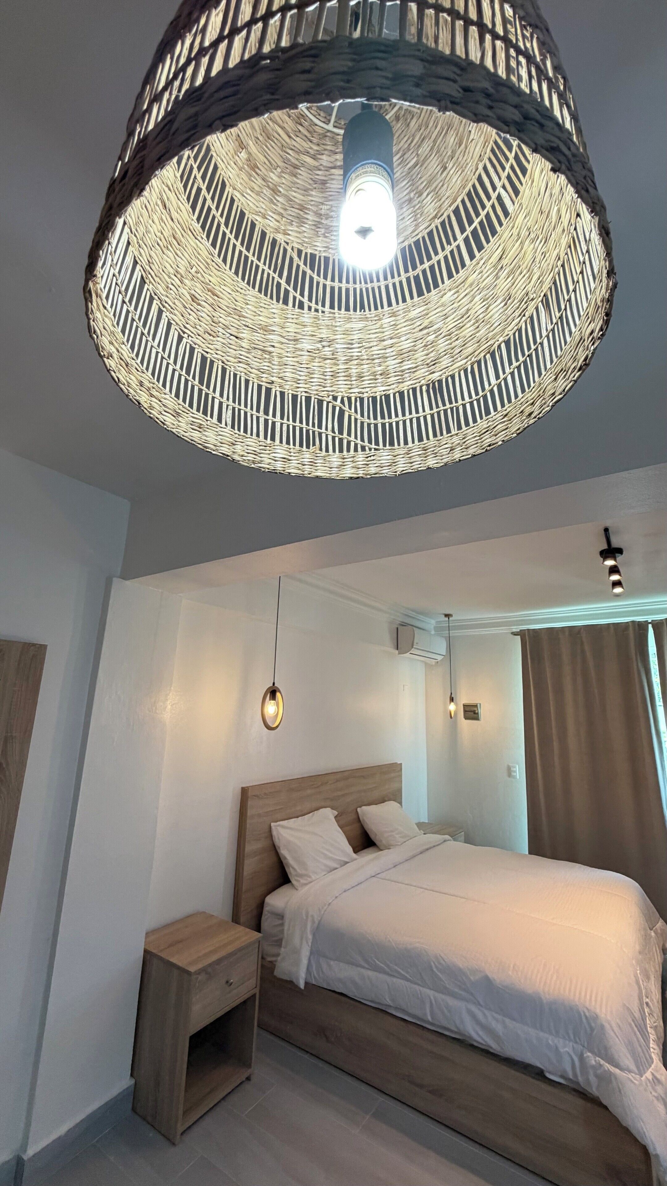 A bedroom with a large bed, a nightstand, a wooden headboard, a woven ceiling light fixture, and a wall-mounted air conditioning unit.