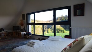 A bedroom with a large window, a bed with white linens, a desk with a chair, and a view of a green landscape.