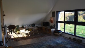 A spacious room with a large window, a flat-screen TV, a dining table, and a tripod lamp.