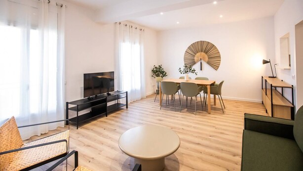 A modern living room with a flat-screen TV, a dining area with a round table and chairs, and a large decorative fan on the wall. - 63IND1125- Brand new apartment (Barcelona)