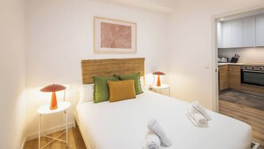 A hotel room with a bed, two bedside tables, a headboard, a kitchen area visible through an open door, and a framed artwork on the wall. - 63IND1125- Brand new apartment (Barcelona)