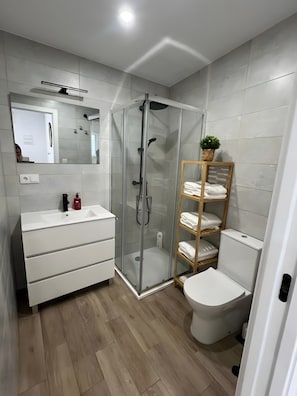 A modern bathroom with a glass-enclosed shower, a white sink with a mirror, and a wooden shelf with towels. - Apartment 'Bajo De Guía - Bajo A' with Wi-Fi and Air Conditioning (Sanlúcar de Barrameda)
