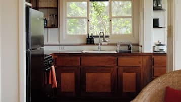 Une cuisine avec des meubles en bois, un réfrigérateur noir, une fenêtre donnant sur un espace verdoyant et un coussin rouge sur un canapé.