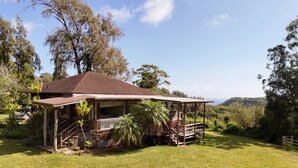 A rustic wooden house with a porch, surrounded by lush greenery and a clear view of the hills.