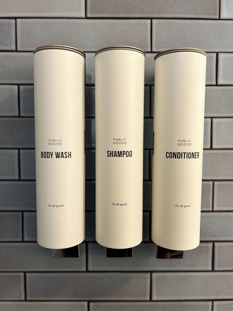 Tres recipients de productes de bany etiquetats com 'BODY WASH', 'XAMPO' i 'CONDITIONER' penjats a la paret.