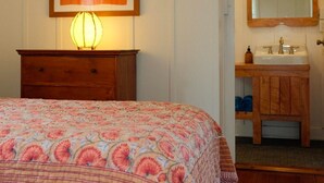Un dormitori amb un llit, una taula de nit amb una làmpada, una obra d'art emmarcada a la paret, i un bany amb un mirall, un lavabo i un armari de fusta.