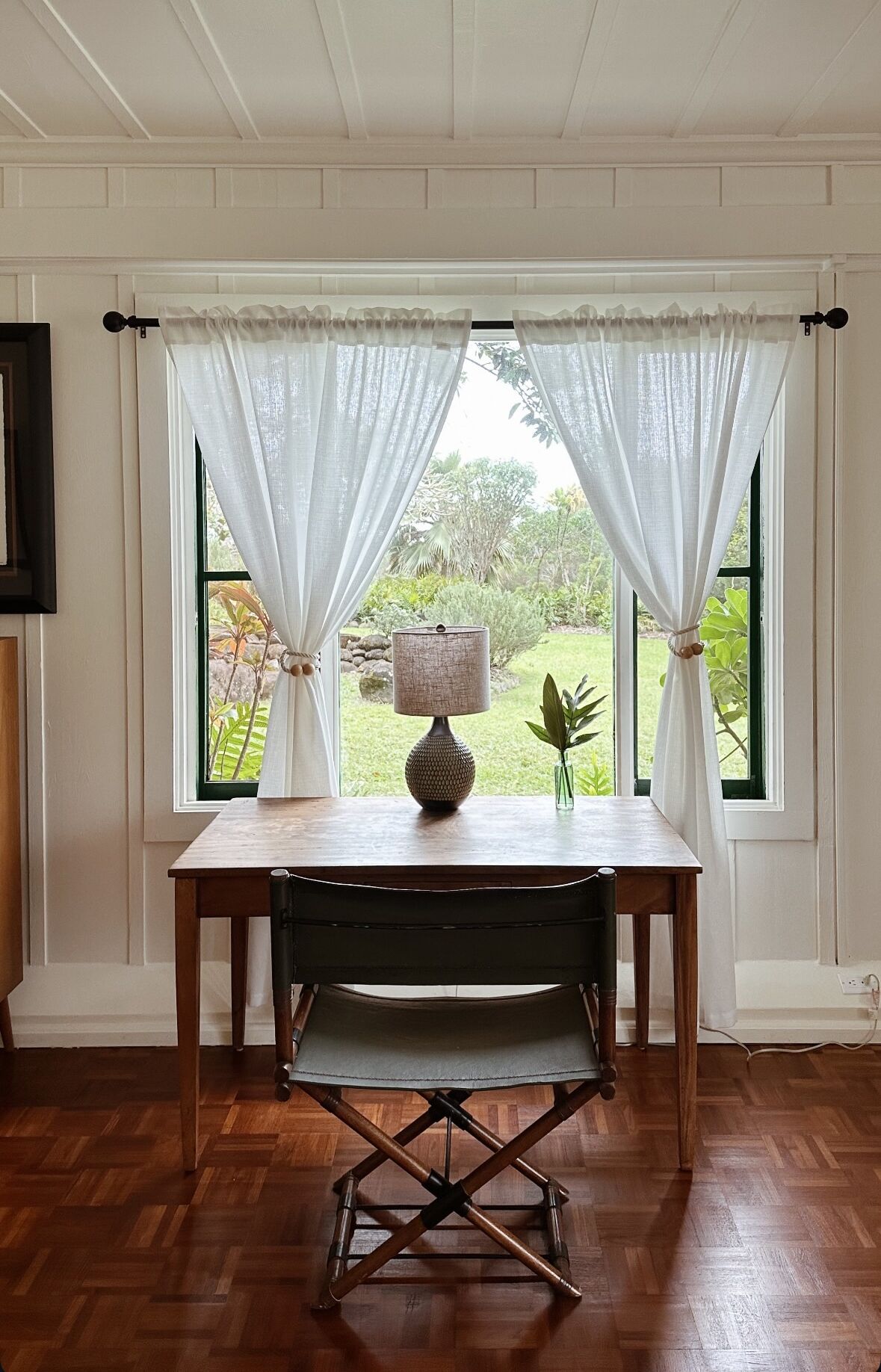 A dining area with a wooden table and a bench, a lamp, and a view of greenery through two windows with white curtains.