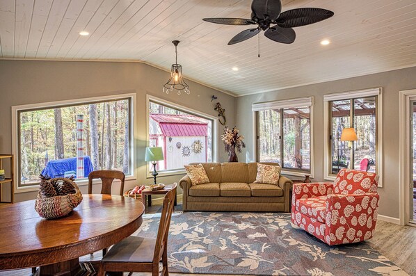 A living room with a wooden ceiling, a ceiling fan, a sofa, a red patterned chair, a dining table, and large windows with a view of trees.