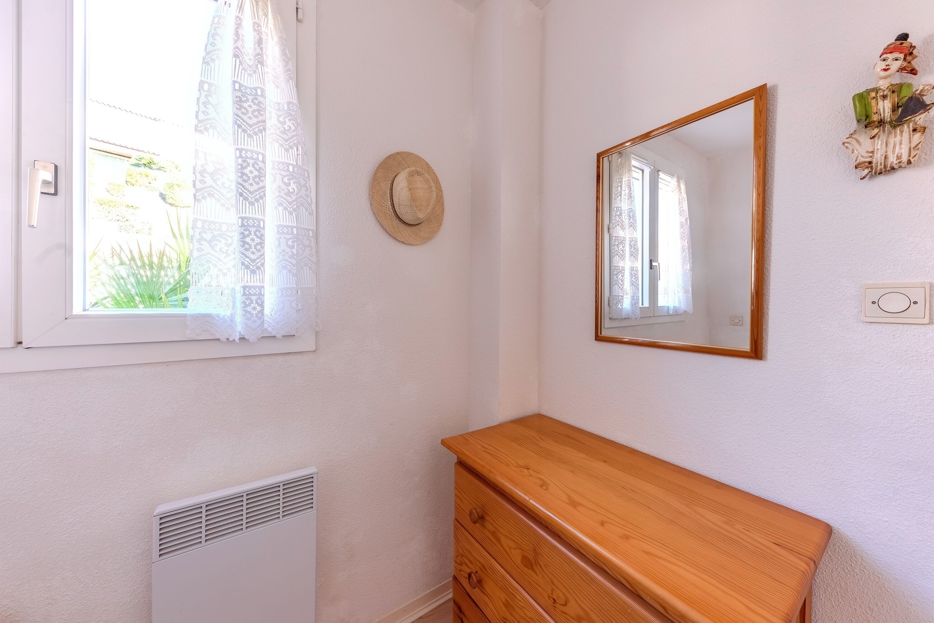 A room with a wooden dresser, a mirror, a window with a patterned curtain, and a wall-mounted hat.