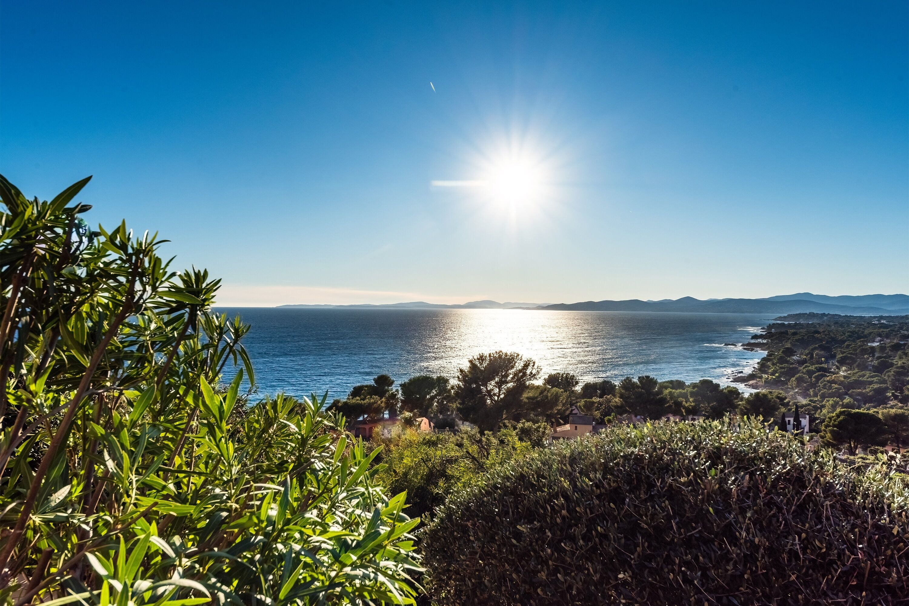 Eine Küstenlandschaft mit blauem Himmel, strahlender Sonne und Blick auf das Meer und ferne Hügel.