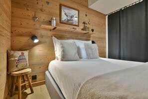 A cozy room with a wooden ceiling, a bed with white bedding, a small stool with a winter-themed pillow, and a framed picture on the wall. - Lys Martagon - Hermine 2 (Le Grand Bornand)