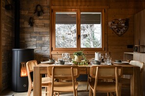 A wooden dining area with a table set for four, a black heater, and a window with a view of trees. - Lys Martagon - Hermine 2 (Le Grand Bornand)