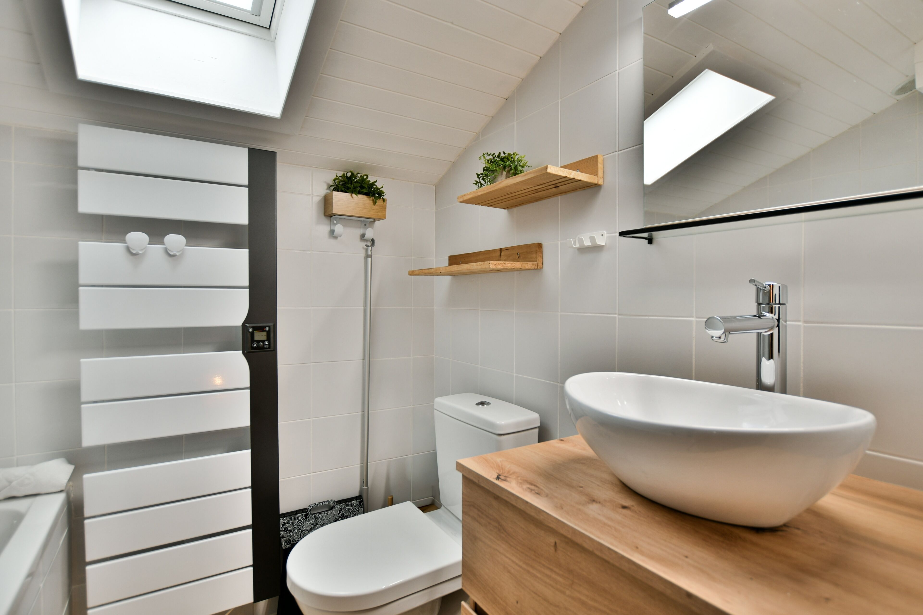 Een moderne badkamer met een wit bad, een wit toilet, een houten wastafelmeubel met een grote witte wasbak en houten planken met planten.