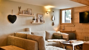 Ein gemütliches Wohnzimmer mit einer Couch, einem Couchtisch und einem an der Wand befestigten Fernseher. Regale mit Dekorationen und ein Fenster, das natürliches Licht hereinlässt.
