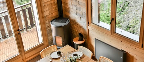 A dining area with a table set for four, a wood-burning stove, and large windows offering a view of the outdoors.