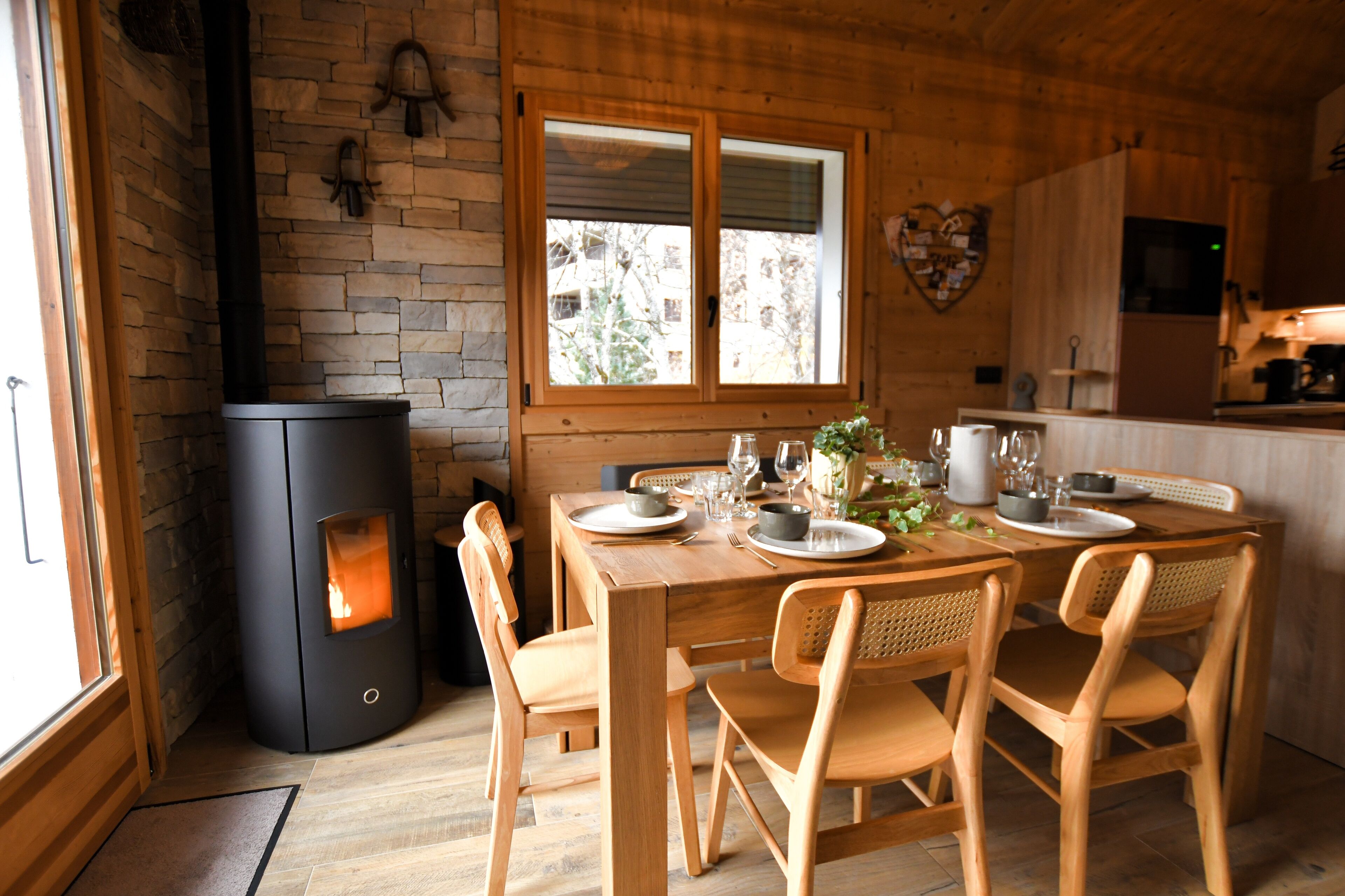 A wooden dining table with chairs, a black wood stove, and a window with a view of buildings.