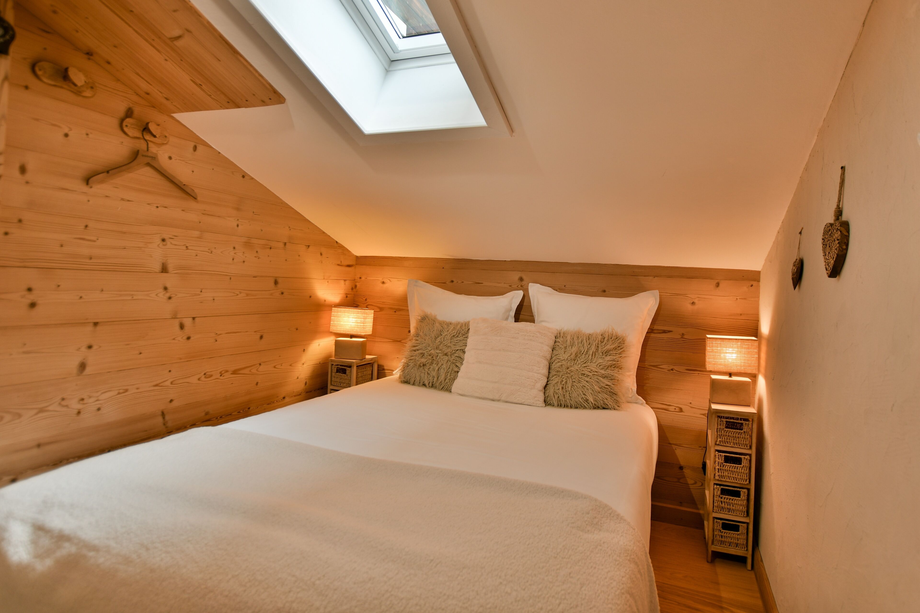 Ein Schlafzimmer mit Dachfenster, Holzwänden, einem Bett mit weißer Bettwäsche, zwei Nachttischen mit Lampen und einer kleinen Holzkommode.