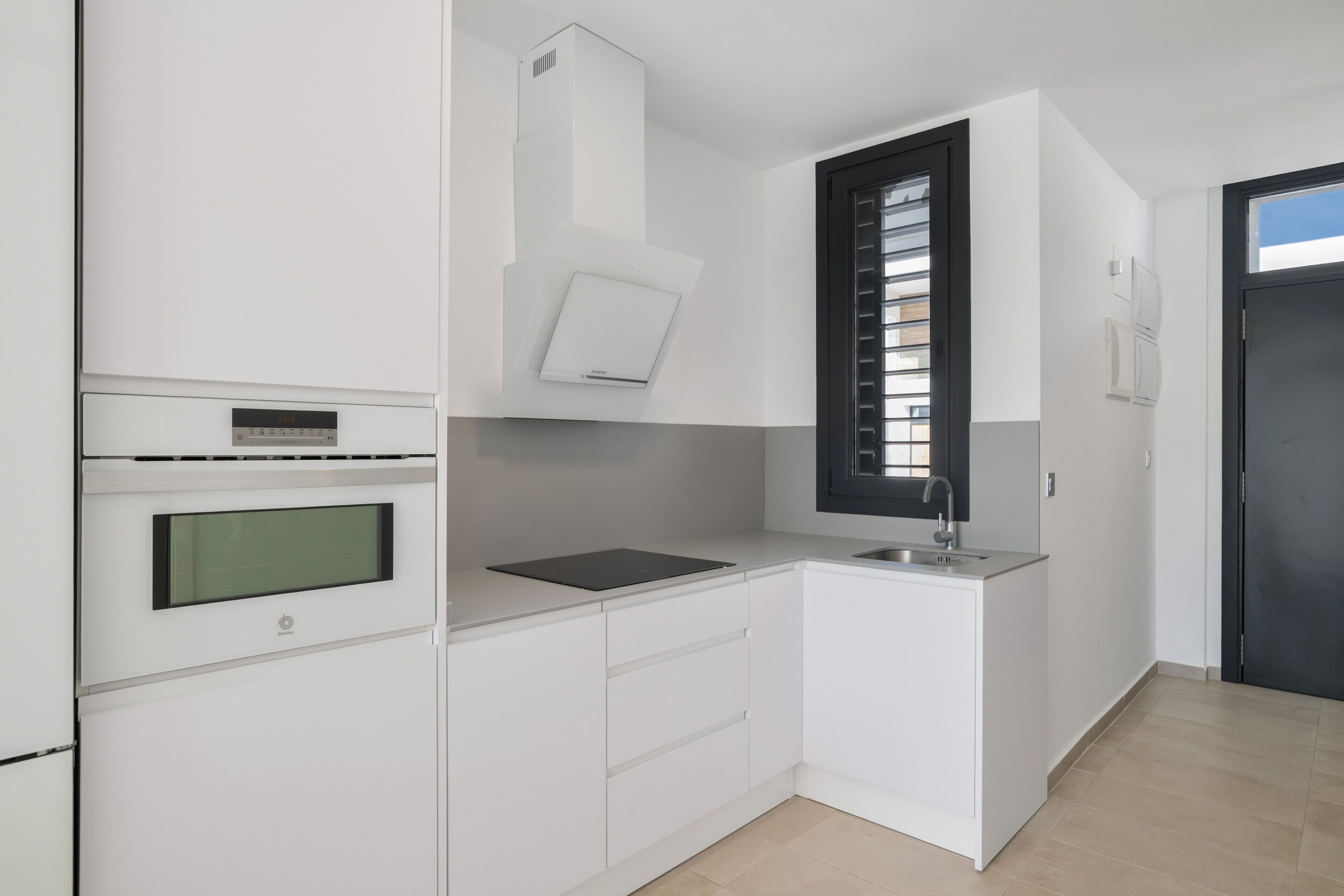 Een moderne keuken met witte kasten, een ingebouwde oven, een raam met zonwering en een deur met een zwarte omlijsting.