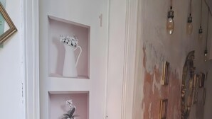 Een gang met een witte deur, planken met vazen, en een muur met afbladderende verf en hangende lampen.
