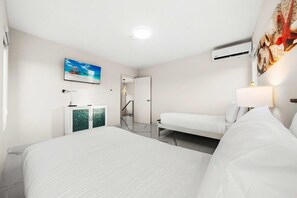 Ein Hotelzimmer mit einem großen Bett, einem kleineren Bett, einem Schreibtisch mit Lampe, einem an der Wand montierten Fernseher und einer Klimaanlage.