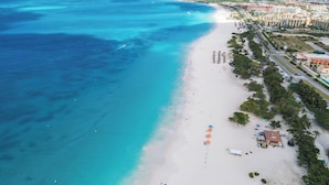 Eine Vogelperspektive zeigt einen Strand mit deutlicher Abgrenzung zwischen dem Meer und dem Sandstrand, eine Reihe von Gebäuden parallel zum Strand sowie einige Sonnenschirme, die Strandmöglichkeiten signalisieren.