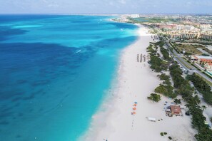 An aerial view of a beach with a clear demarcation between the ocean and the sandy shore, a row of buildings parallel to the beach, and a few umbrellas indicating beachside amenities. - Prime location • Private pool & BBQ (Noord)