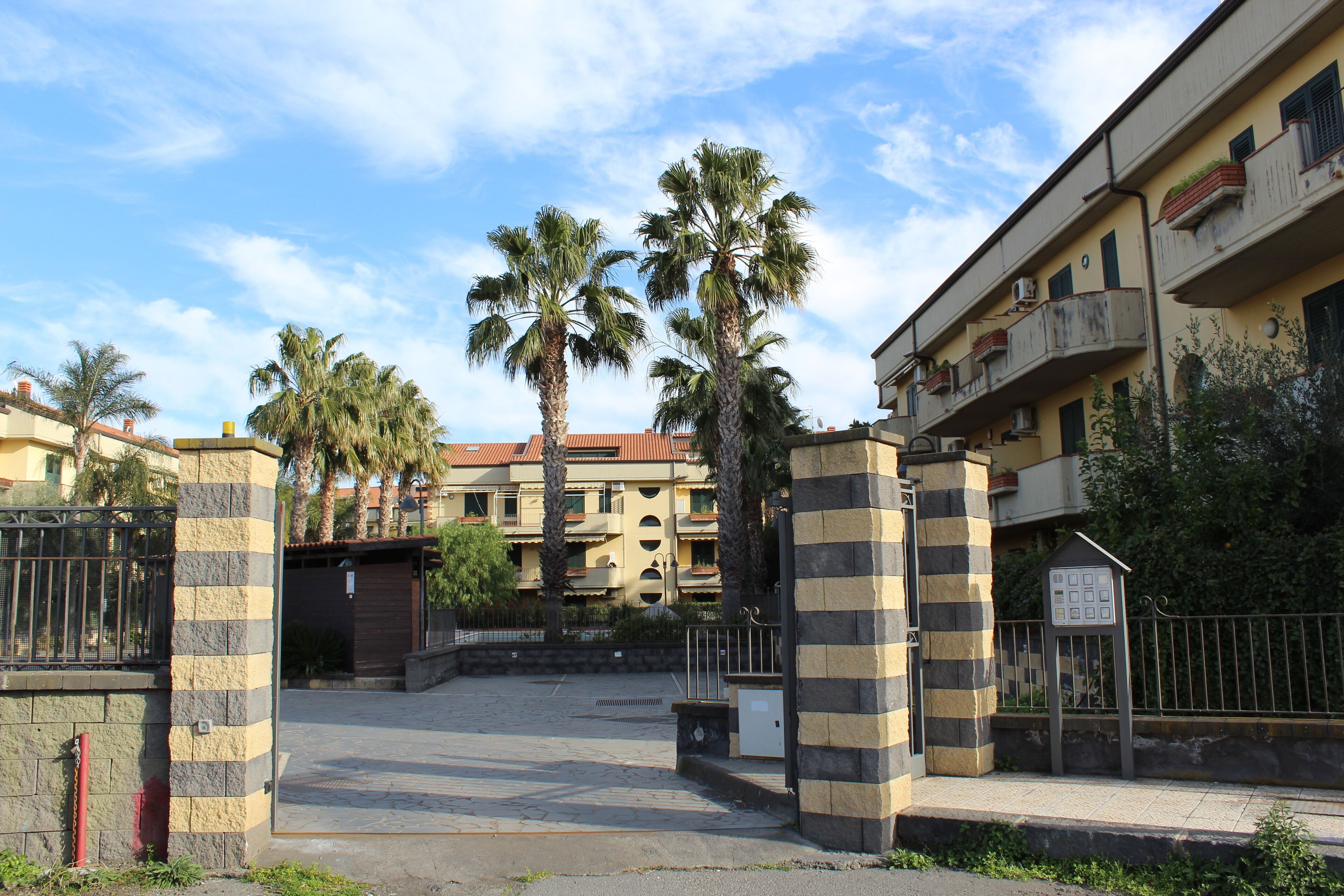 Un ingresso recintato con palme, un muro in pietra e un edificio con balconi.