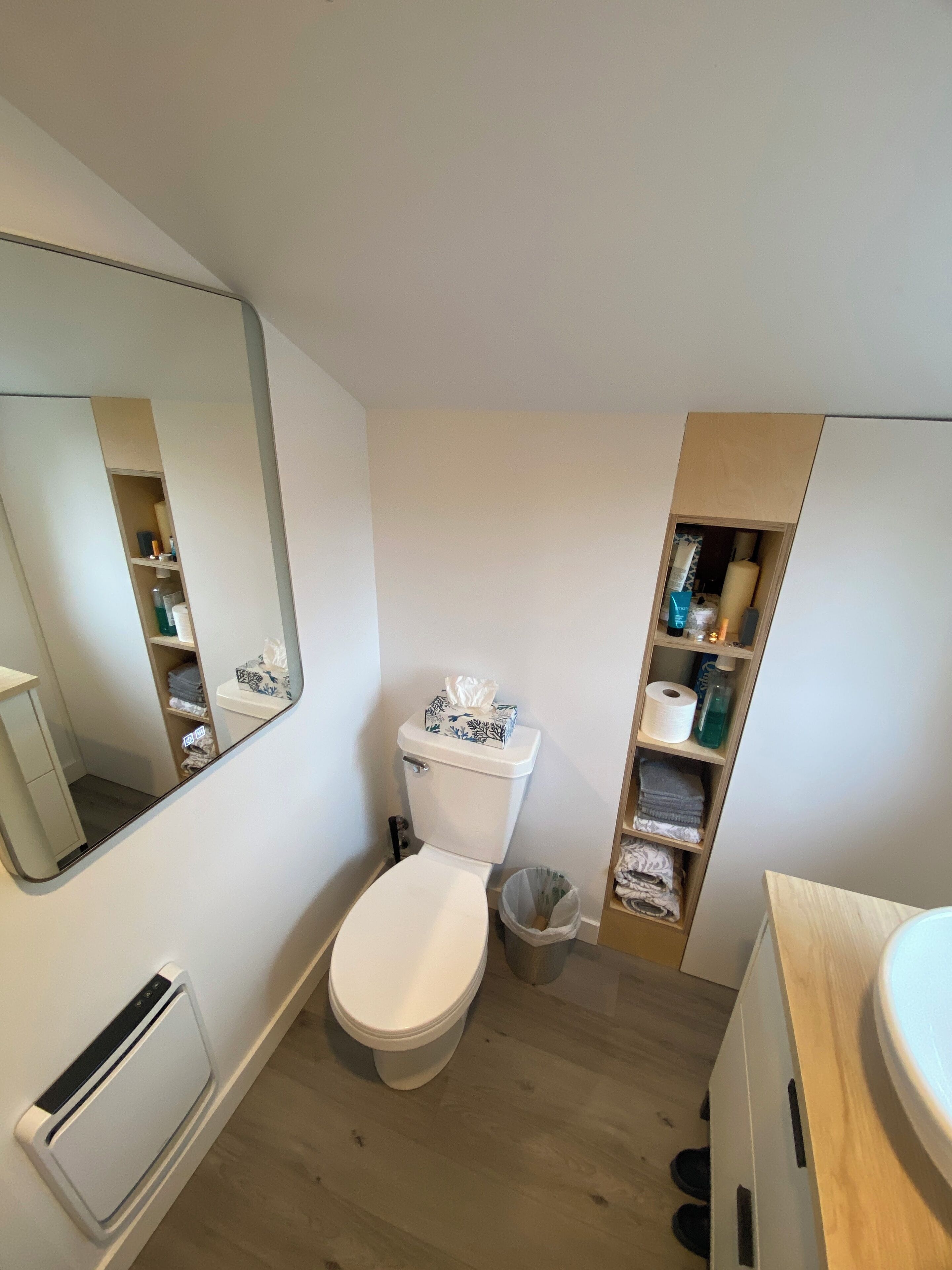 A modern bathroom with a toilet, a mirror, a shelf with toiletries, and a trash bin.