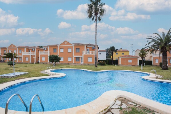 Una piscina con agua azul cristalina, rodeada por una terraza de piedra y contigua a una fila de edificios anaranjados con balcones.