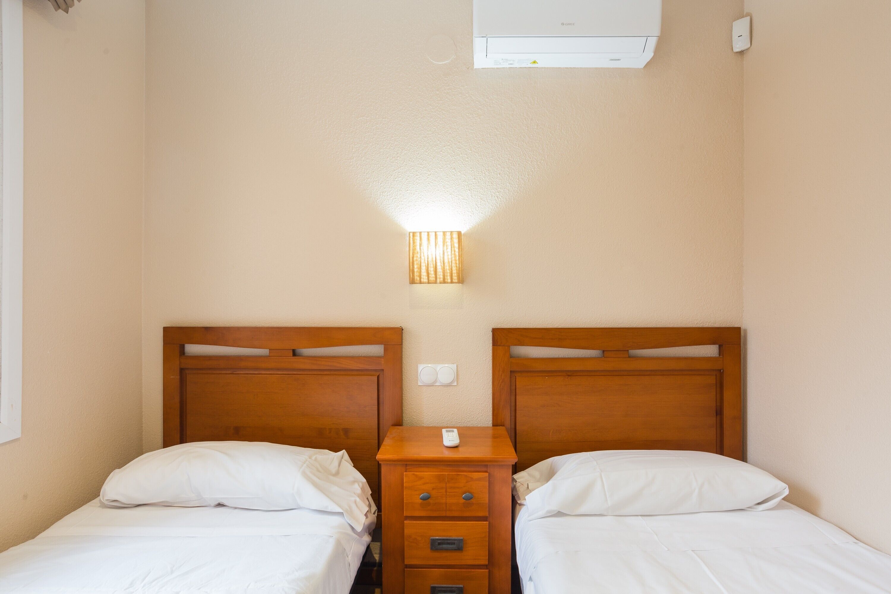 Ein Hotelzimmer mit zwei Einzelbetten, einem Nachttisch aus Holz mit Lampe und einer Wand-Klimaanlage.