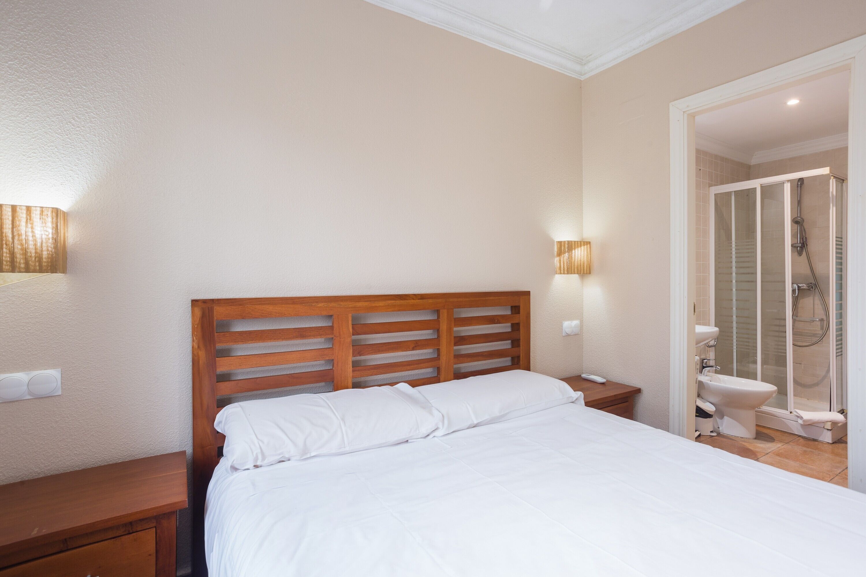 Une chambre à coucher avec une tête de lit en bois, un lit avec une literie blanche, une table de chevet, une lampe fixée au mur et une salle de bain avec une douche et des toilettes.