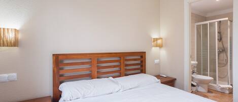 Une chambre à coucher avec une tête de lit en bois, un lit avec une literie blanche, une table de chevet, une lampe fixée au mur et une salle de bain avec une douche et des toilettes.