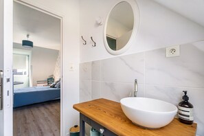 Een badkamer met een houten wastafelmeubel, een witte wastafel, een spiegel en uitzicht op een slaapkamer.