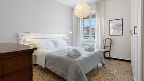 Hotellihuone, jossa on sänky, puinen lipasto, tuoli, työpöytä, riippuvalaisin ja ikkuna, jossa on verhot.