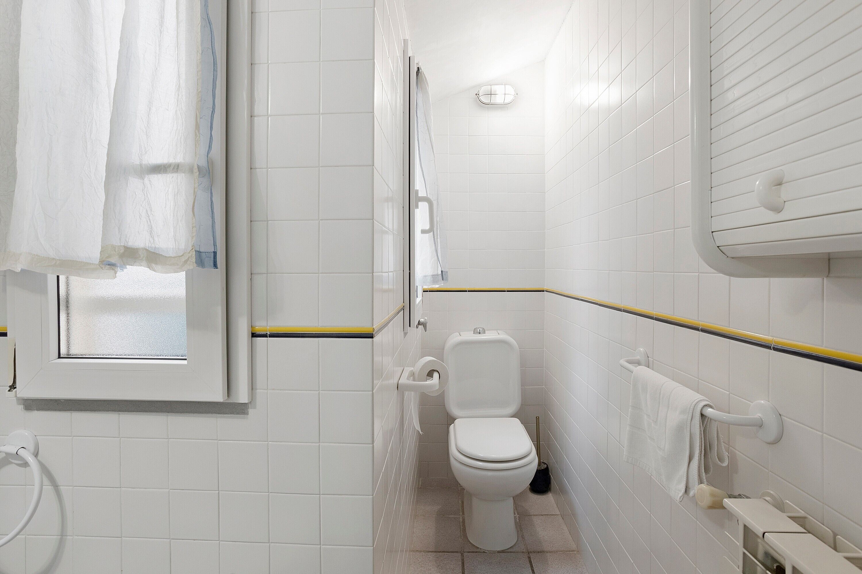 Une salle de bain avec des carreaux blancs, des toilettes, un porte-serviettes et une fenêtre avec un rideau.