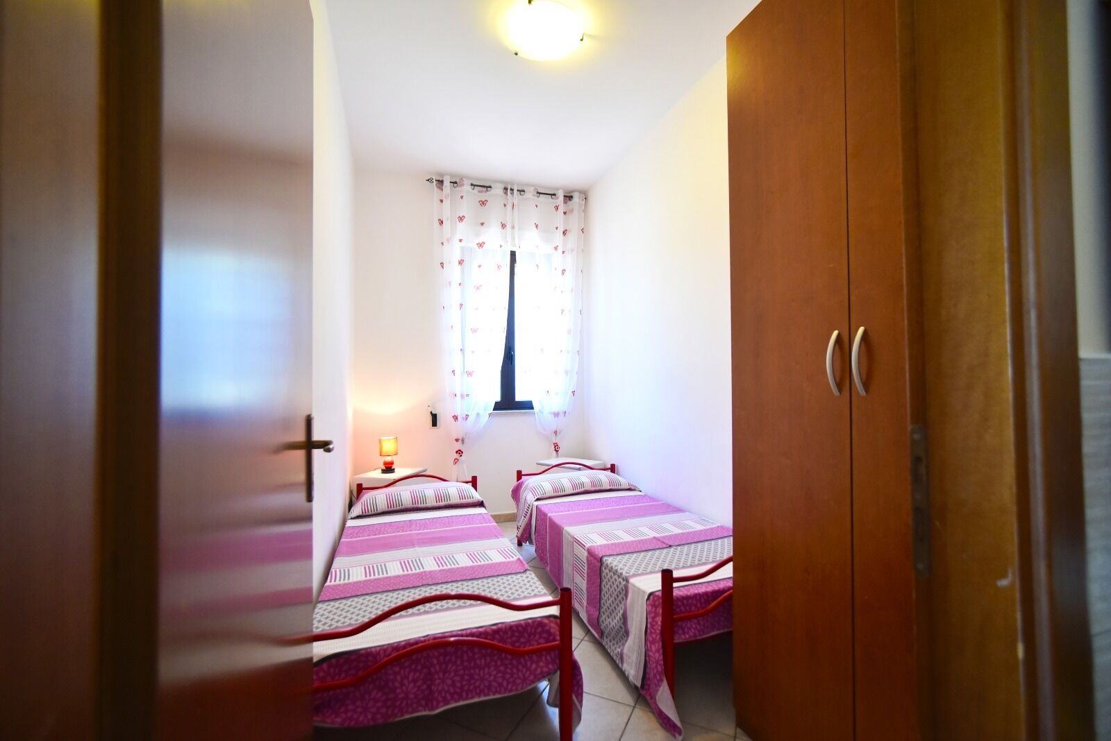 Un dormitorio con dos camas, una ventana con cortinas, una mesita de noche con lámpara y un armario.