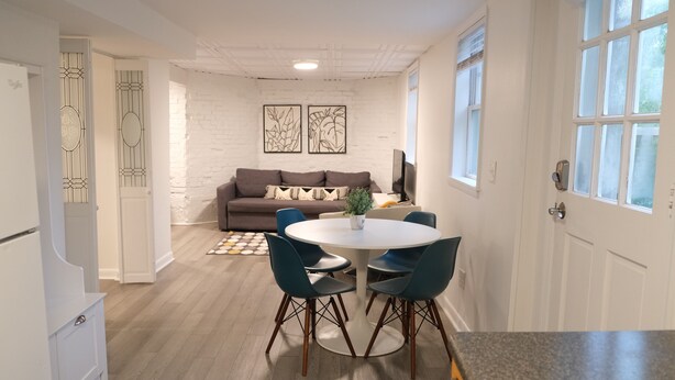 A modern living room with a white brick wall, a brown sofa, and a round dining table with four chairs. - Charming Getaway! Heart of Asheville Suite (Asheville)
