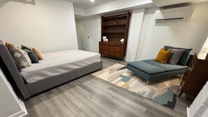 A modern bedroom with a large bed, a sofa, a wooden dresser, and a white brick accent wall. - Charming Getaway! Heart of Asheville Suite (Asheville)