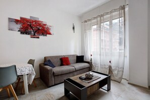 Ein Wohnzimmer mit einer Couch, einem Couchtisch, einem Esstisch mit Stühlen und einem großen Fenster mit durchsichtigen Vorhängen.
