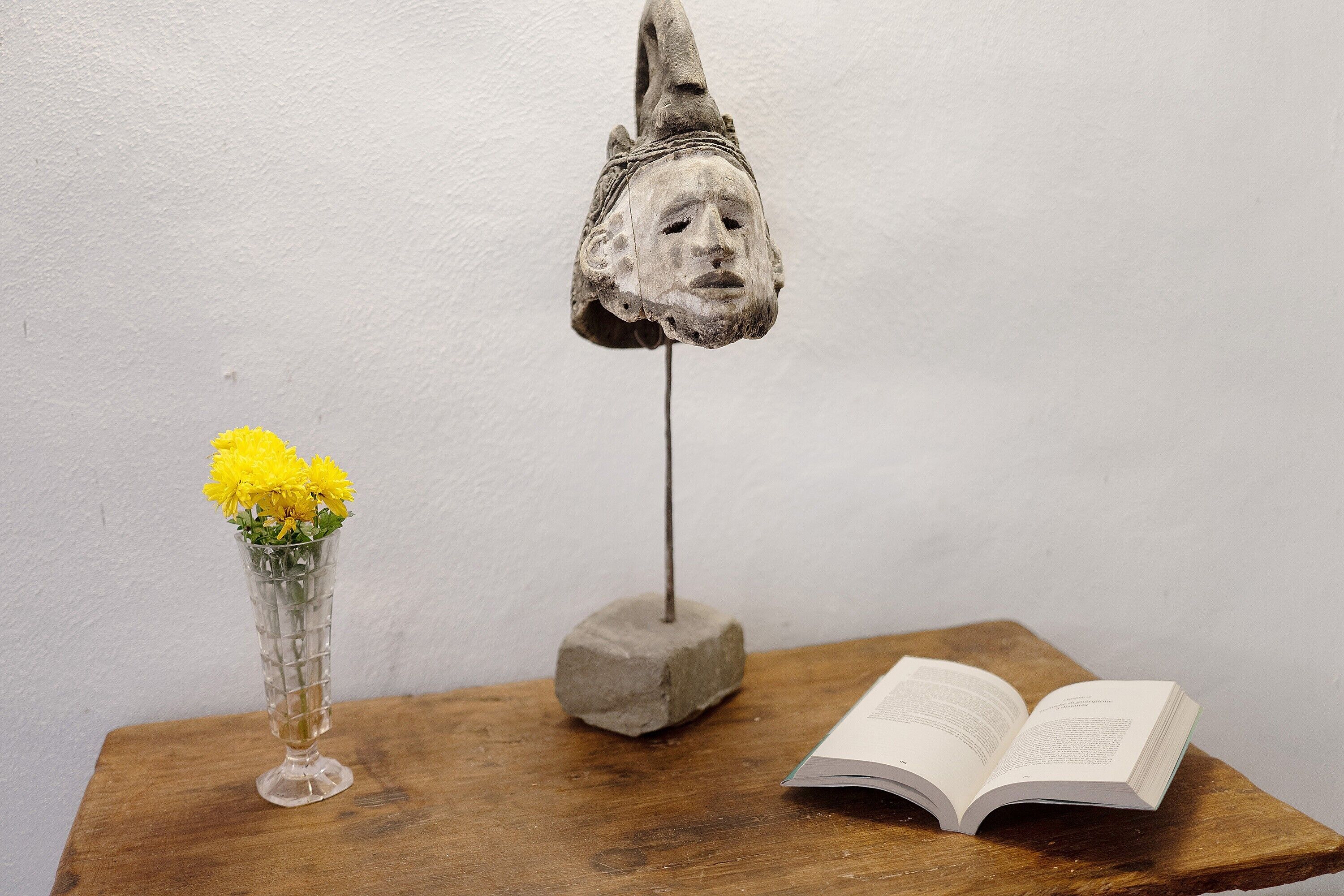Een houten tafel met een vaas gele bloemen, een boek en een stenen beeldhouwwerk.