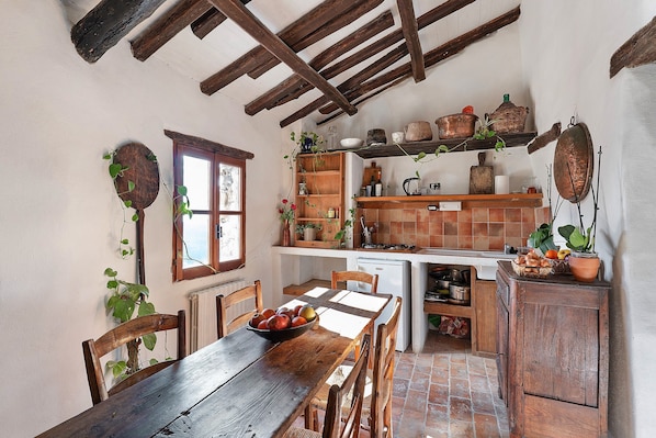 A rustic kitchen with wooden beams, a dining table with chairs, and a variety of kitchenware and plants.