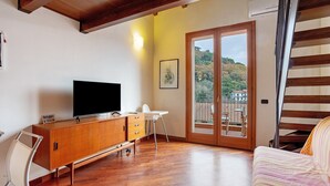 Una habitación con un armario de madera, un televisor, una silla blanca, un sofá y vistas a una ladera a través de un ventanal.