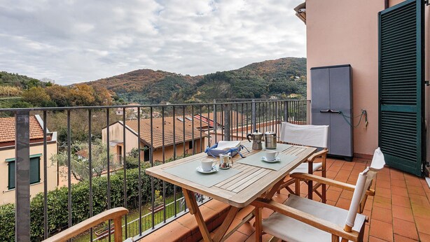 Un balcón con una mesa dispuesta para dos, vista del paisaje circundante y un edificio con un persiano verde.