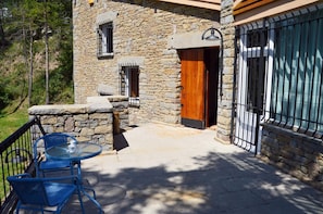 A stone building with a wooden door, a balcony with blue chairs, and a glass table.