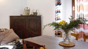 Een houten eettafel met een geweven tafelonderzetter, een glazen vaas met gele bloemen en een houten kast op de achtergrond.