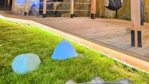 Une terrasse en bois agrémentée d’un hamac et de guirlandes lumineuses, entourée de rochers et d’herbe.