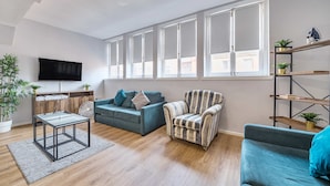 A modern living room with a sofa, armchair, and a glass coffee table. There are windows with white frames, a flat-screen TV, and a wooden floor. - Host Stay Apartment 8 Stanley Street (Liverpool)