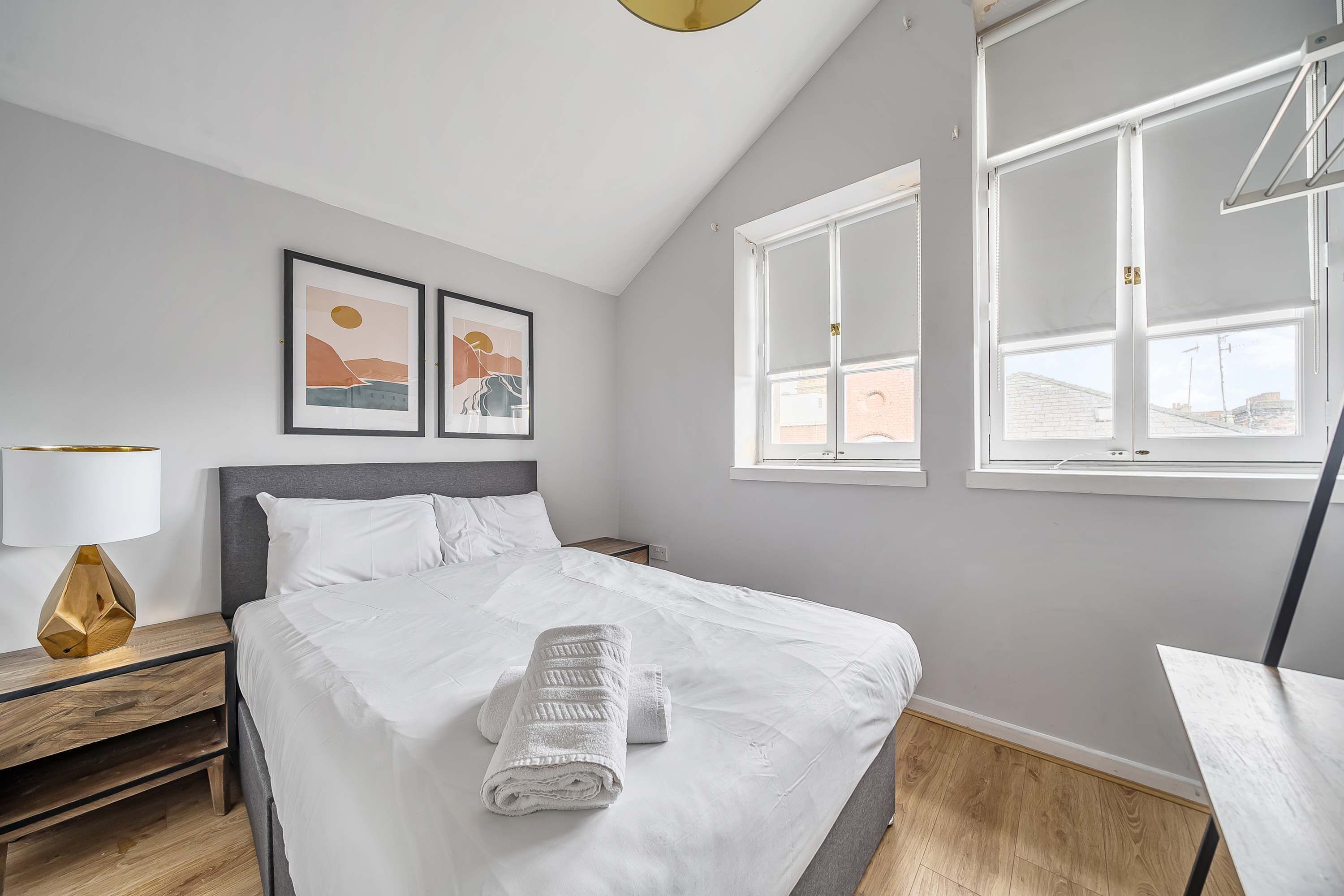 A neatly made bed with white linens and a folded towel, a bedside lamp, and two framed artworks on the wall.