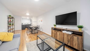 Una sala de estar moderna con sofá, una mesa de centro de vidrio, un televisor sobre un mueble de madera, una zona de comedor con sillas y una mesa, y una cocina al fondo.